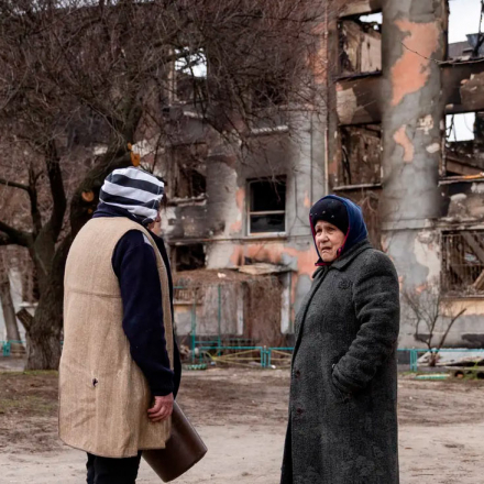 Two women talk in front of a destroyed building in Schastia