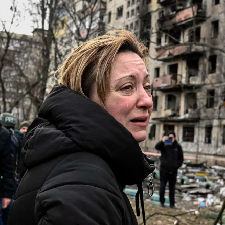 A woman reacts as she stands outside a ruined apartment building